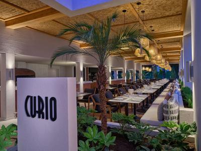 Elegantes Hotelrestaurant mit Holzdach, Palmen und langen Tischen für Gäste.