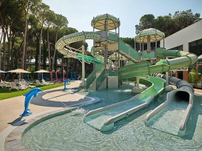 Wasserrutsche mit mehreren Bahnen in einem Hotelpool, umgeben von Sonnenliegen und Bäumen.