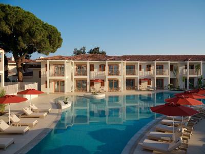 Moderner Hotelpool mit Liegen und roten Sonnenschirmen vor zweistöckigem Gebäude bei klarem Himmel.