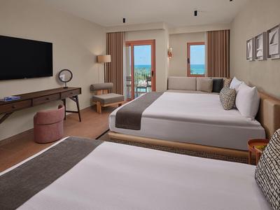 Helles Hotelzimmer mit zwei Doppelbetten, Schreibtisch, Sesseln und Blick auf das Meer.