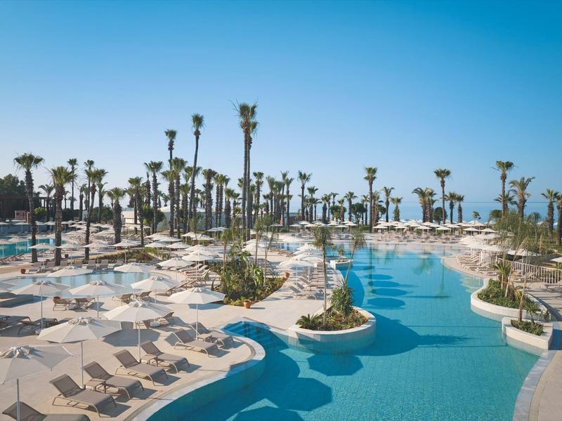 Großer Poolbereich mit Palmen und Liegestühlen unter blauem Himmel in einem Resort.