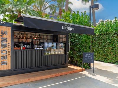 Kleiner schwarzer Kiosk mit "Brown's" Schild, Palmen im Hintergrund und grünem Sichtschutz.