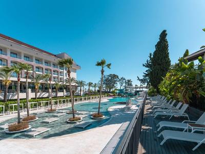 Blick auf einen Pool mit Palmen, Liegestühlen und einem mehrstöckigen Hotel bei sonnigem Himmel.