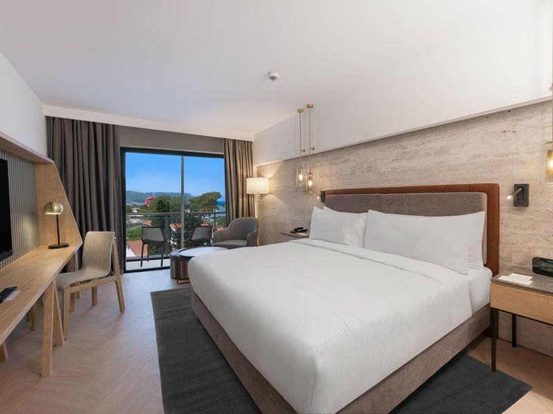 Modernes Hotelzimmer mit großem Bett, Balkon, Schreibtisch, Fernseher und minimalistischer Einrichtung.