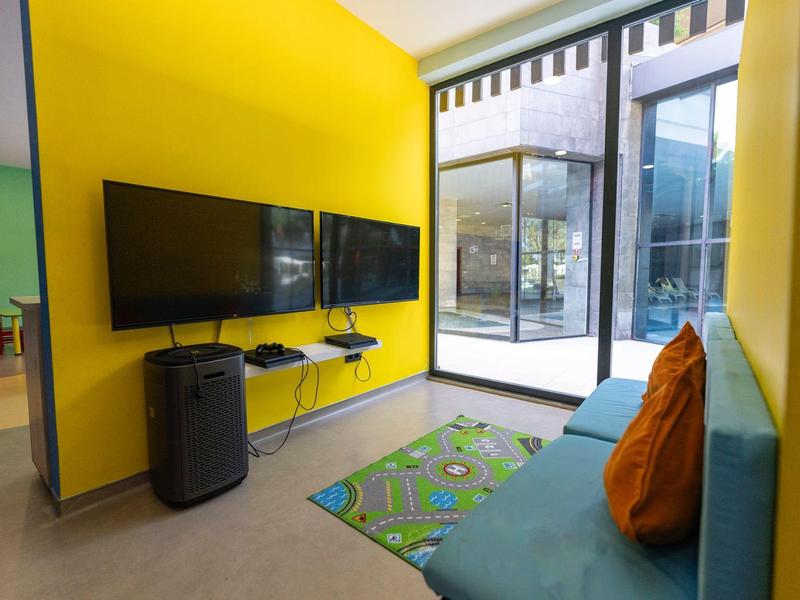 Modernes Wartezimmer mit gelben Wänden, blauer Couch, orangen Kissen und zwei Fernsehern an der Wand.