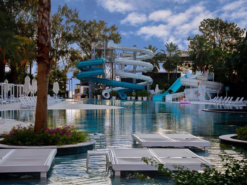 Freizeitbad mit Wasserrutschen, Sitzgelegenheiten und Palmen unter bewölktem Himmel.