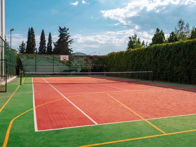 Leerer Tennisplatz mit rotem Spielfeld, grünem Rand und von Bäumen umgebenem Zaun bei blauem Himmel.