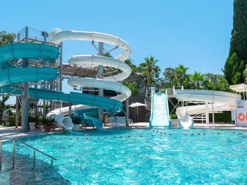 Großer Außenpool mit mehreren Wasserrutschen und umgeben von Palmen und blauen Himmel.