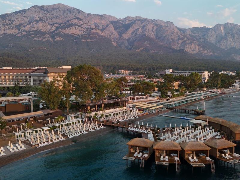 Küstenresort mit langem Steg, Liegestühlen am Wasser und Bergen im Hintergrund bei klarem Himmel.