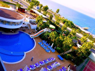 Blick auf Pool mit Wasserrutsche, blauen Liegestühlen, grünen Palmen und Meer im Hintergrund.
