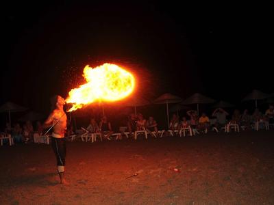 Person bläst eine große Feuersäule in dunkler Nacht am Strand mit Stühlen im Hintergrund.