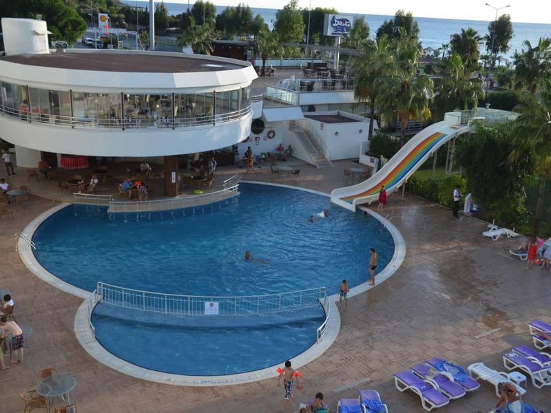 Großer runder Pool mit Wasserrutsche und umliegender Terrasse, daneben ein zweistöckiges Gebäude.