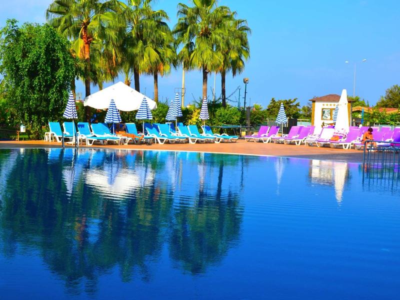 Blauer Pool mit Liegestühlen, Sonnenschirmen und Palmen bei klarem Himmel und Sonnenschein.