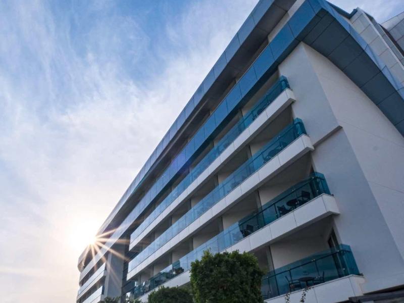 Vista de un edificio de hotel de varios pisos con balcones y autos frente bajo cielo soleado.