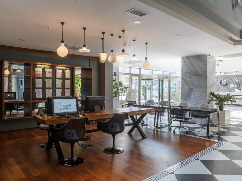 Moderna zona de salón de hotel con suelos de madera y mármol, mesas y sillas, iluminada por lámparas colgantes.