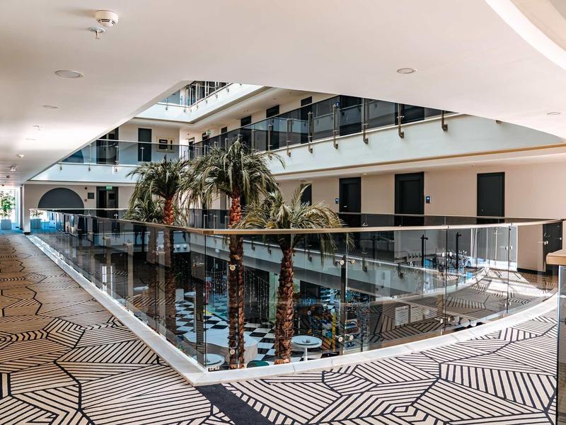 Pasillo luminoso de hotel con baldosas geométricas modernas y barandilla de vidrio, decorado con palmeras.