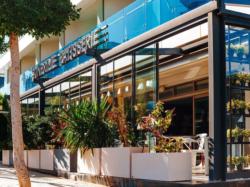 Vista exterior de un hotel moderno con paredes de vidrio azul y plantas frente a la entrada.