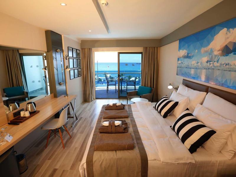 Habitación de hotel moderna con cama doble, escritorio y vista al mar a través de grandes ventanas.