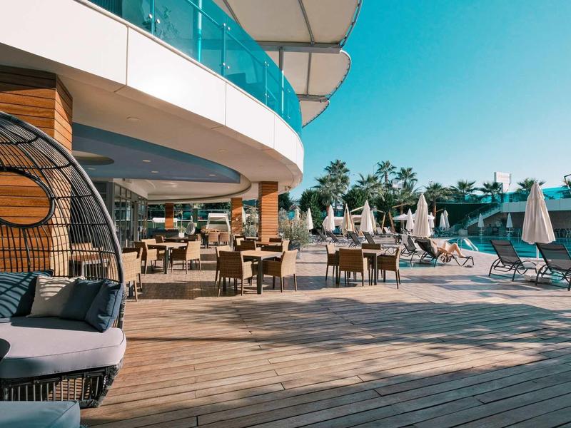 Terraza con suelo de madera, mesas, sillas y área de piscina bajo cielo azul en un resort hotelero.