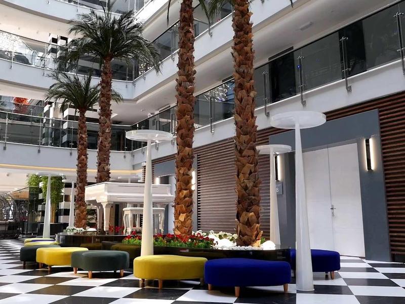 Vestíbulo de hotel moderno y luminoso con palmeras, bancos coloridos y suelo a cuadros en blanco y negro.