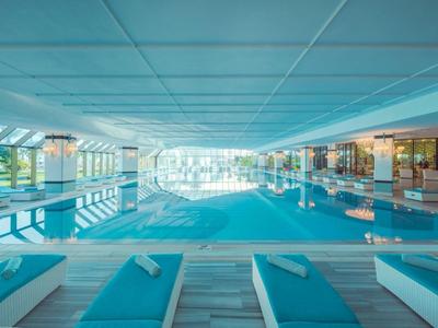 Piscina cubierta luminosa con tumbonas y grandes ventanales que muestran árboles y área exterior.