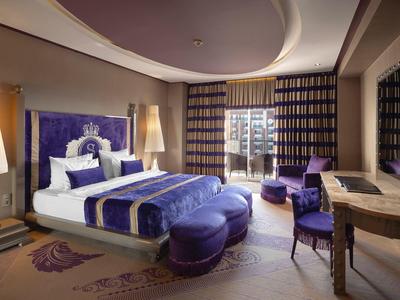 Habitación de hotel elegante con acentos en púrpura, cama doble, asientos y grandes ventanas con cortinas.