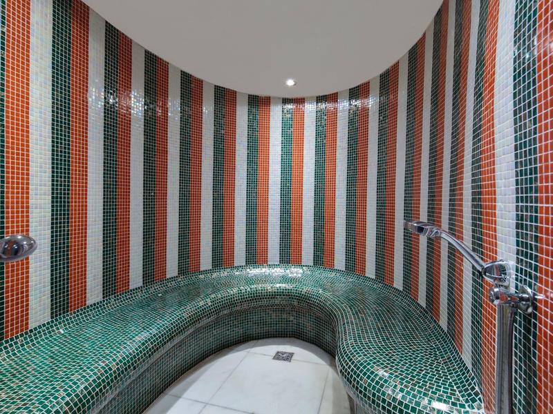 Sala de vapor redonda con banco verde y paredes de azulejos rayados en naranja y verde.