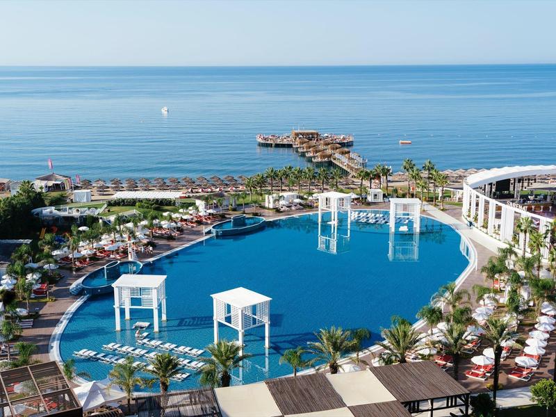 Gran piscina del hotel con estructuras blancas junto a palmeras y vista al mar bajo cielo despejado.