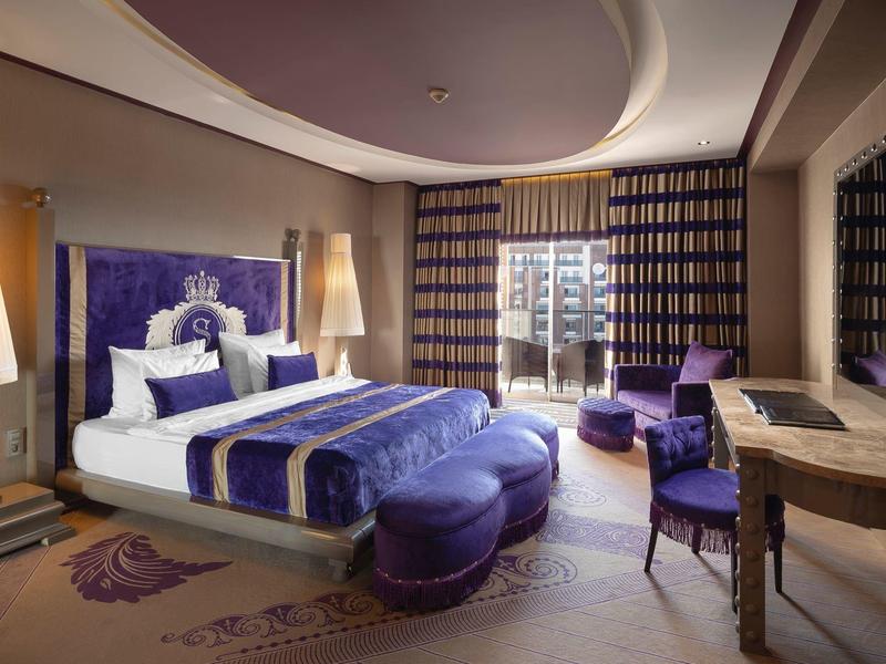 Habitación de hotel elegante con acentos en púrpura, cama doble, asientos y grandes ventanas con cortinas.