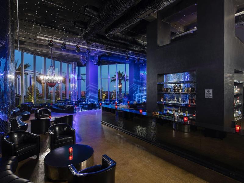 Área de bar de hotel moderna con sillones de cuero negro, barra de madera y luz ambiental azul por la noche.