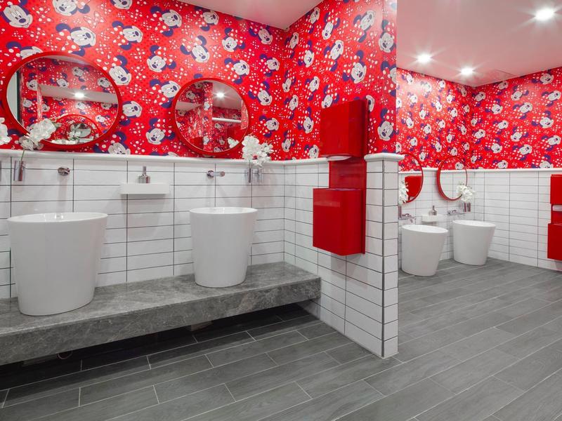 Baño moderno de hotel con sanitarios blancos y azulejos rojos con flores en la pared.