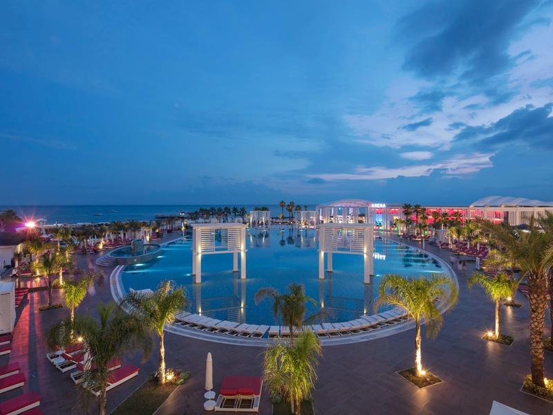 Vista nocturna de una gran piscina iluminada en un hotel con palmeras y zonas de descanso.