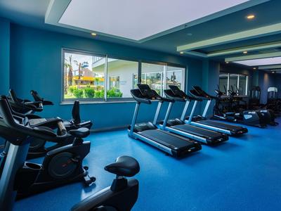 Moderní posilovna s běžeckými pásy a rotopedy, modrá podlaha a velká okna s výhledem do zahrady.