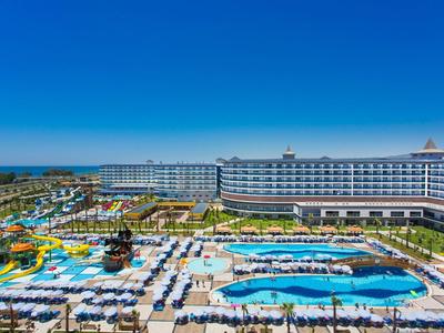 Pohled na moderní hotel s bazény, skluzavkami a řadami slunečníků pod jasnou oblohou.