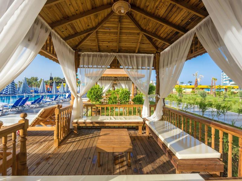 Dřevěná pergola s bílými závěsy a lavicemi v rekreačním středisku.