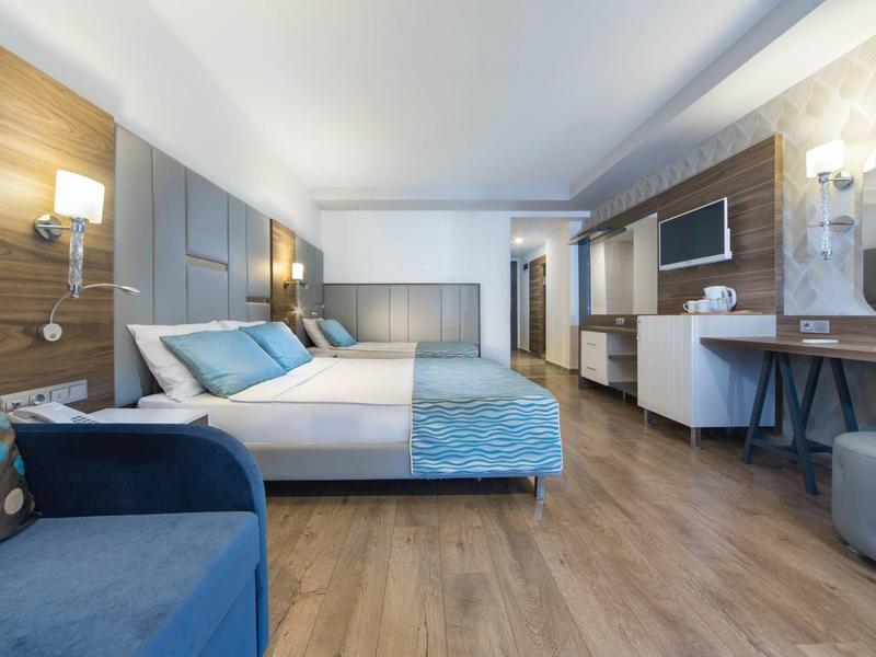 Moderní hotelový pokoj s manželskou postelí, modrým gaučem a světlou dřevěnou podlahou.