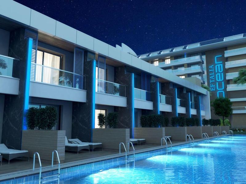 Noční osvětlený bazén před moderním hotelem s balkony a lehátky.