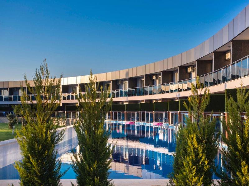 Moderní hotel s bazénem ve tvaru půlměsíce a vegetací pod modrou oblohou.