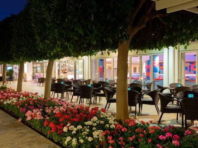 Wieczorny taras z krzesłami i stołami, otoczony kwiatami i drzewami przed oświetlonym budynkiem.