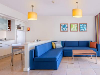 Nowoczesny salon z niebieską narożną sofą, drewnianym stołem i otwartą kuchnią w jasnym pomieszczeniu.