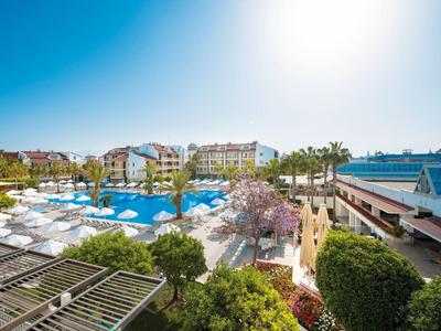 Großer blauer Pool mit weißen Sonnenliegen, umgeben von grünen Bäumen und mehrstöckigen Gebäuden.