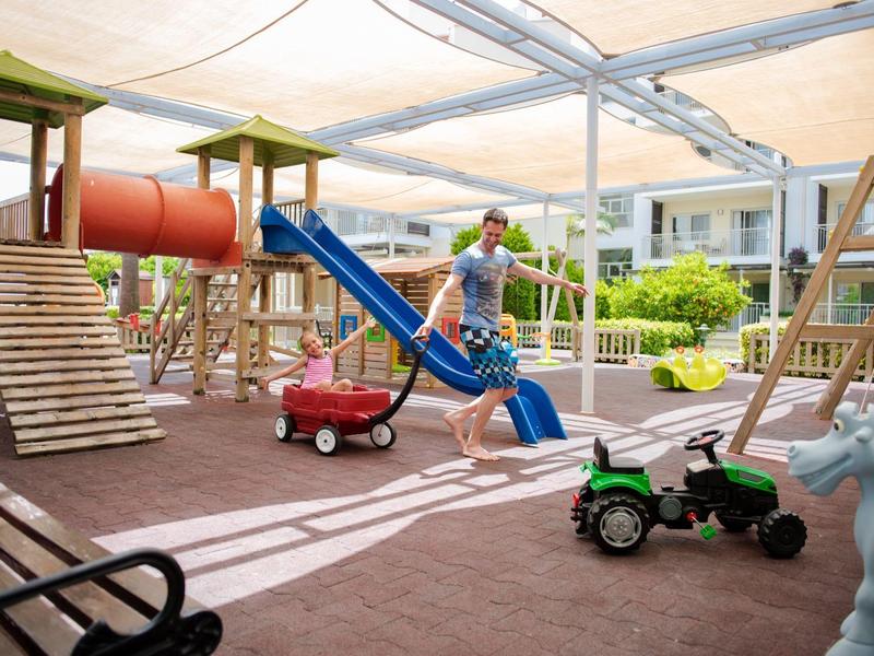 Plac zabaw na świeżym powietrzu z zjeżdżalniami, huśtawkami i zabawkowymi traktorami w ogrodzie hotelowym.