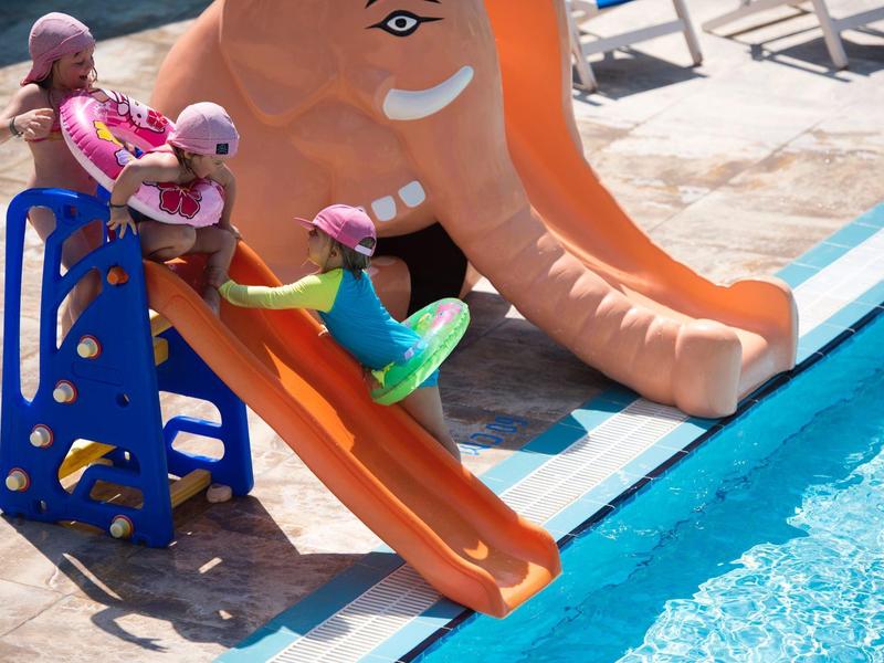 Dzieci zjeżdżają ze zjeżdżalni w kształcie pomarańczowego słonia do basenu w słoneczny dzień.