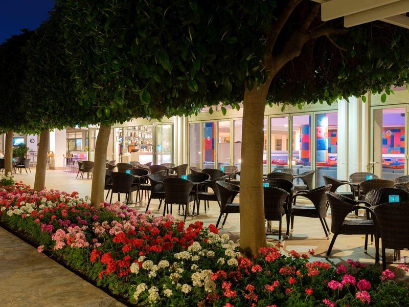 Wieczorny taras z krzesłami i stołami, otoczony kwiatami i drzewami przed oświetlonym budynkiem.