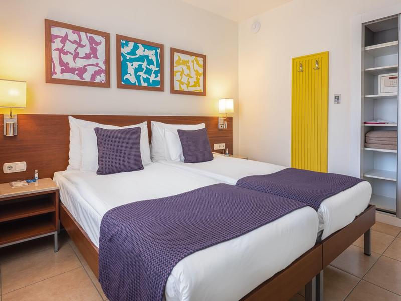 Jasny pokój hotelowy z dwoma pojedynczymi łóżkami i fioletowymi narzutami.