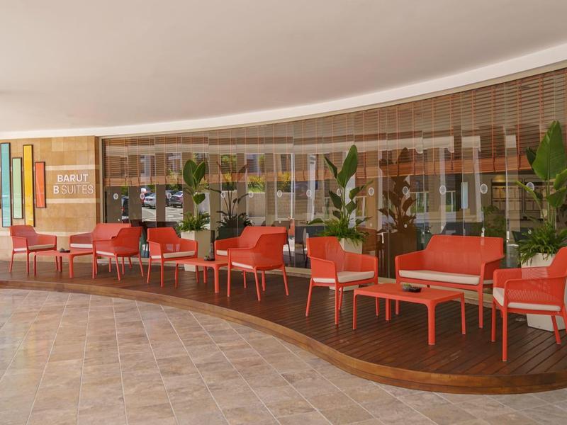 Nowoczesny lobby hotelowy z czerwonymi fotelami, stolikami i dużymi roślinami przy ścianie szklanej.