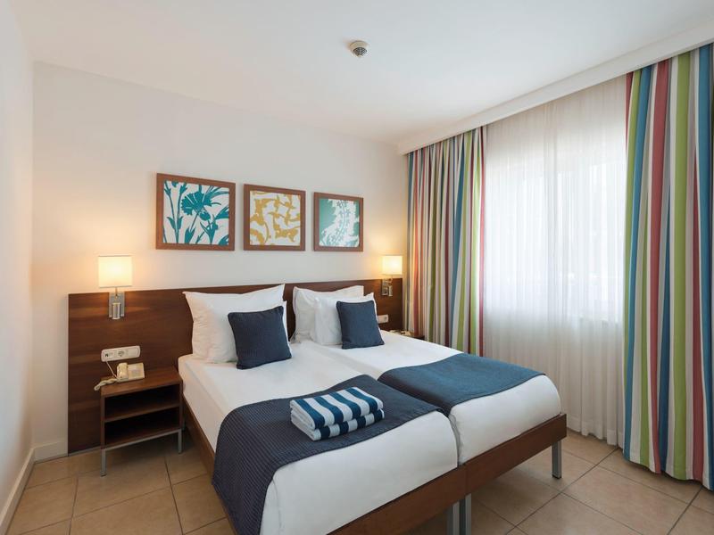 Nowoczesny pokój hotelowy z dwoma pojedynczymi łóżkami, kolorowymi zasłonami i trzema obrazami na ścianie.