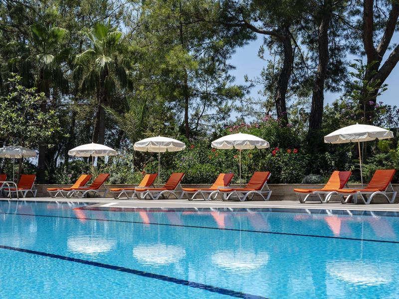 Clear pool with orange lounge chairs and white umbrellas along the edge under trees.