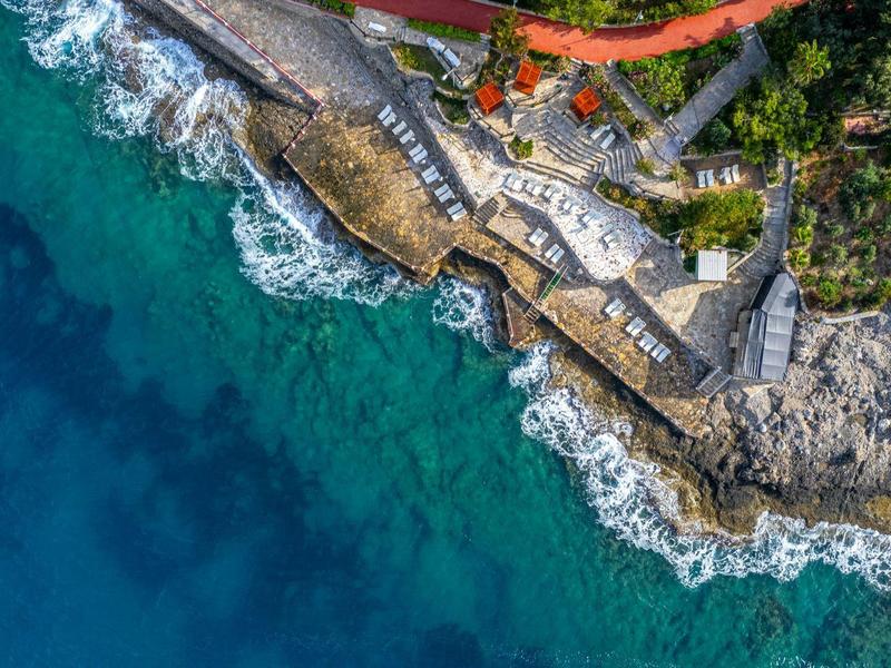 Aerial view of a rocky coastline with clear turquoise water and buildings along the shore.