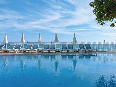 Ein ruhiger Pool mit weißen Liegestühlen und blauen Sonnenschirmen unter wolkigem Himmel.